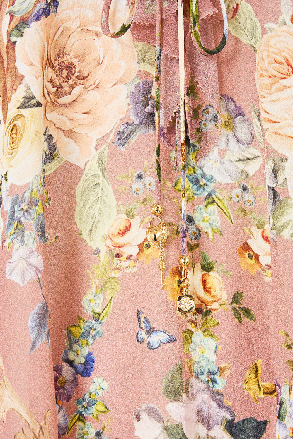 Close-up of a dusty pink floral fabric with large rose motifs, smaller flowers, green leaves, and butterfly details, plus a drawstring with small gold charms.