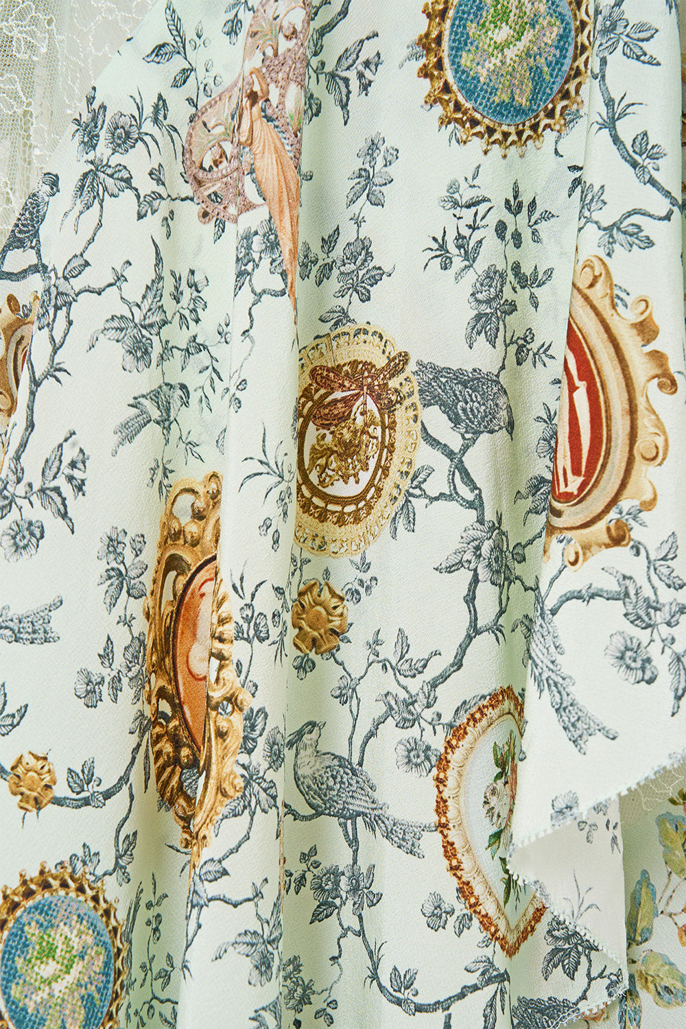 A close-up of a draped fabric featuring a classic toile pattern with blue birds, floral motifs, and ornate gold and red medallions on a pale green background.